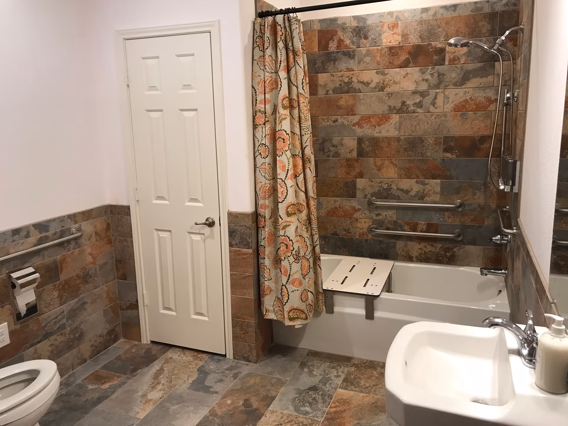 A bathroom with a white toilet, a white sink with a soap dispenser, and a bathtub with a floral patterned shower curtain. The walls and floor are tiled with multicolored stone tiles. There are two grab bars on the bathtub wall and a shower seat inside the bathtub. A closed white door is visible on the back wall.