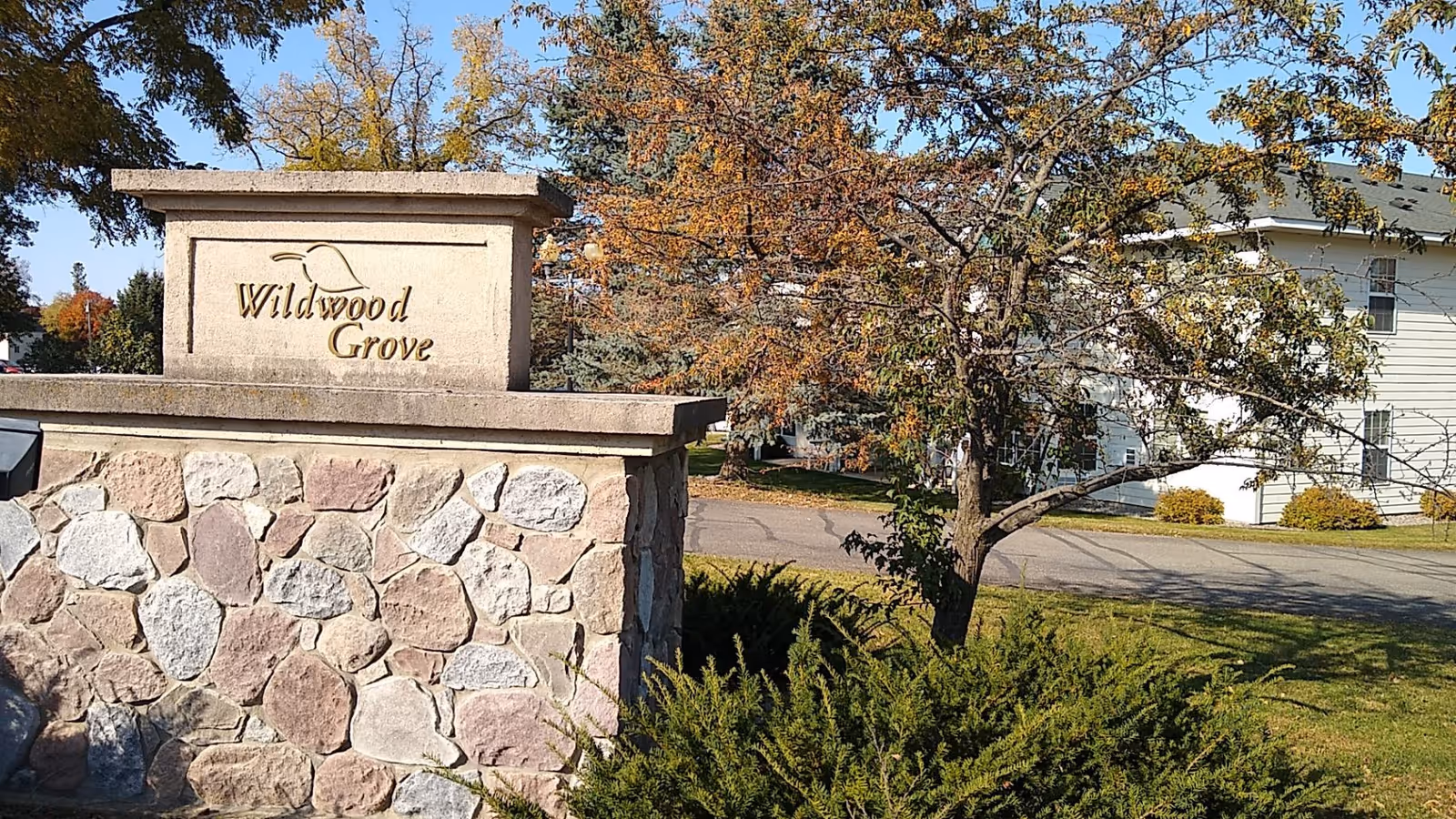 Stone entrance sign reading "Wildwood Grove" beside a tree with a white building and driveway in the background.