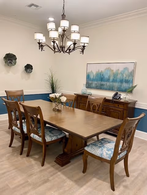 A formal dining room with a wooden table surrounded by six upholstered chairs, a chandelier overhead, a sideboard, and wall art.