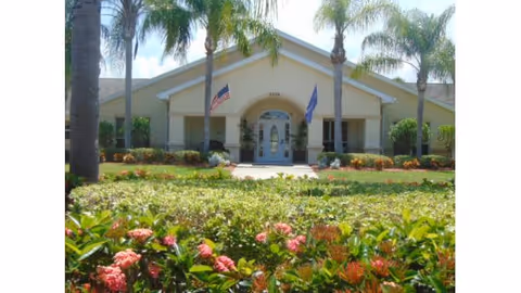 Front exterior view of a single-story building with a beige facade, an arched entrance, and two flags displayed near the doorway. The building is surrounded by palm trees, green shrubs, and flowering plants under a partly cloudy sky.
