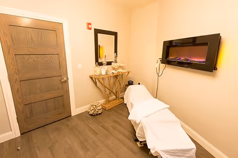 A small, cozy room with a massage table covered in white sheets, a wooden door, a decorative wooden console table with glass jars and a mirror above it, and a modern electric fireplace mounted on the wall.