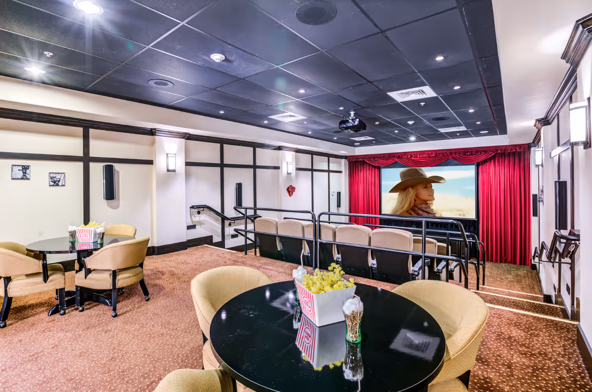 Interior view of a small movie theater room with beige cushioned seats arranged in rows facing a large screen showing a woman in a cowboy hat. The room has a black ceiling with recessed lighting, beige walls with dark wood trim, and carpeted floor. In the foreground, there are round black tables with beige chairs and popcorn containers as decoration.