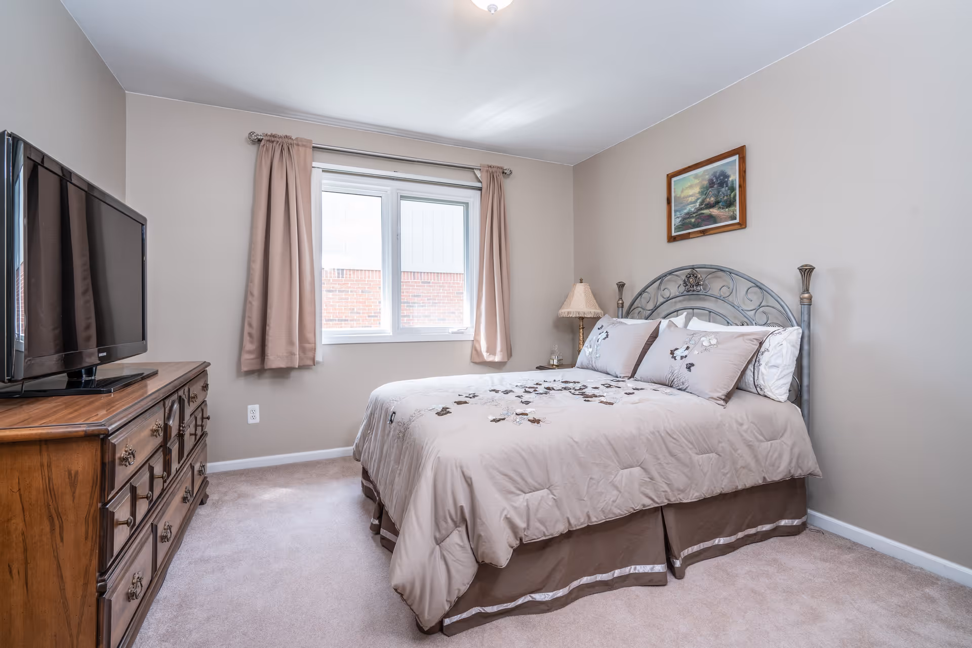 A bedroom with a bed featuring a decorative metal headboard and beige bedding with floral embroidery. There is a wooden dresser with a flat-screen TV on top, a window with beige curtains, a bedside table with a lamp, and a framed painting on the wall.
