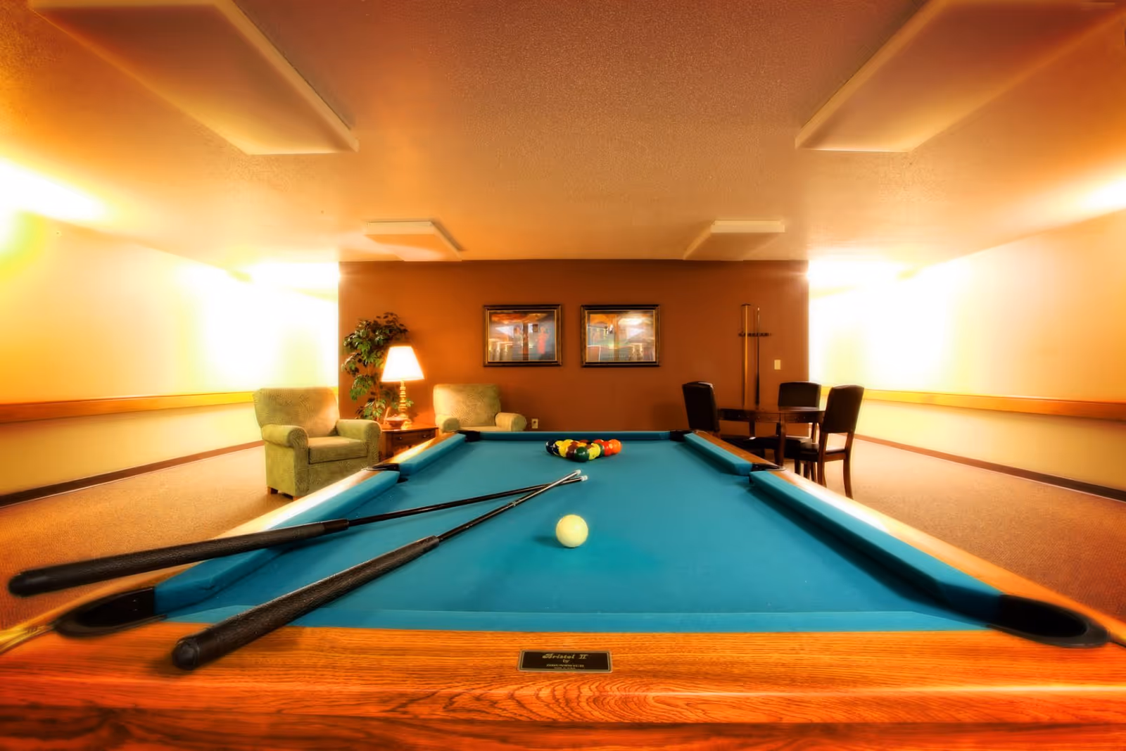 A recreational room featuring a pool table with cues and balls set up for a game. The room has warm lighting, two armchairs with a side table and lamp, a potted plant, and a small table with four chairs against a brown accent wall with two framed pictures.