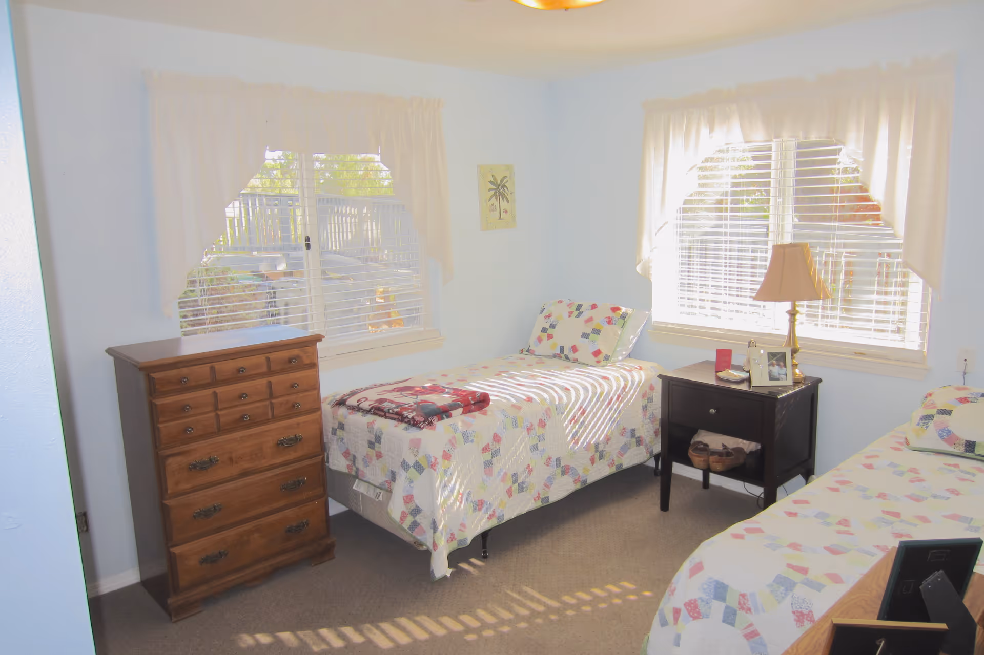 A bright bedroom with two twin beds covered in patchwork quilts. There is a wooden chest of drawers on the left side and a dark wooden nightstand with a lamp, photo frame, and small items between the beds. Two windows with white curtains let in natural light.