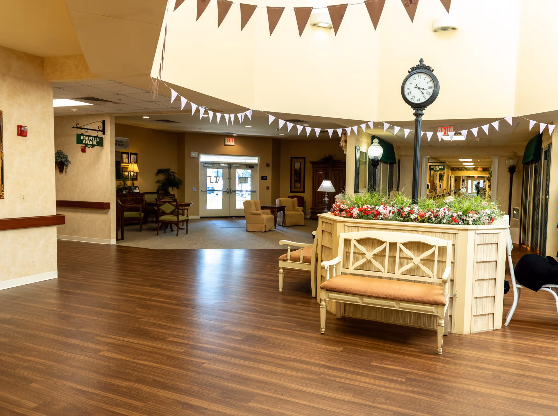Spacious senior living facility lobby with wooden floors, a central planter topped by a clock post, benches and a seating area.
