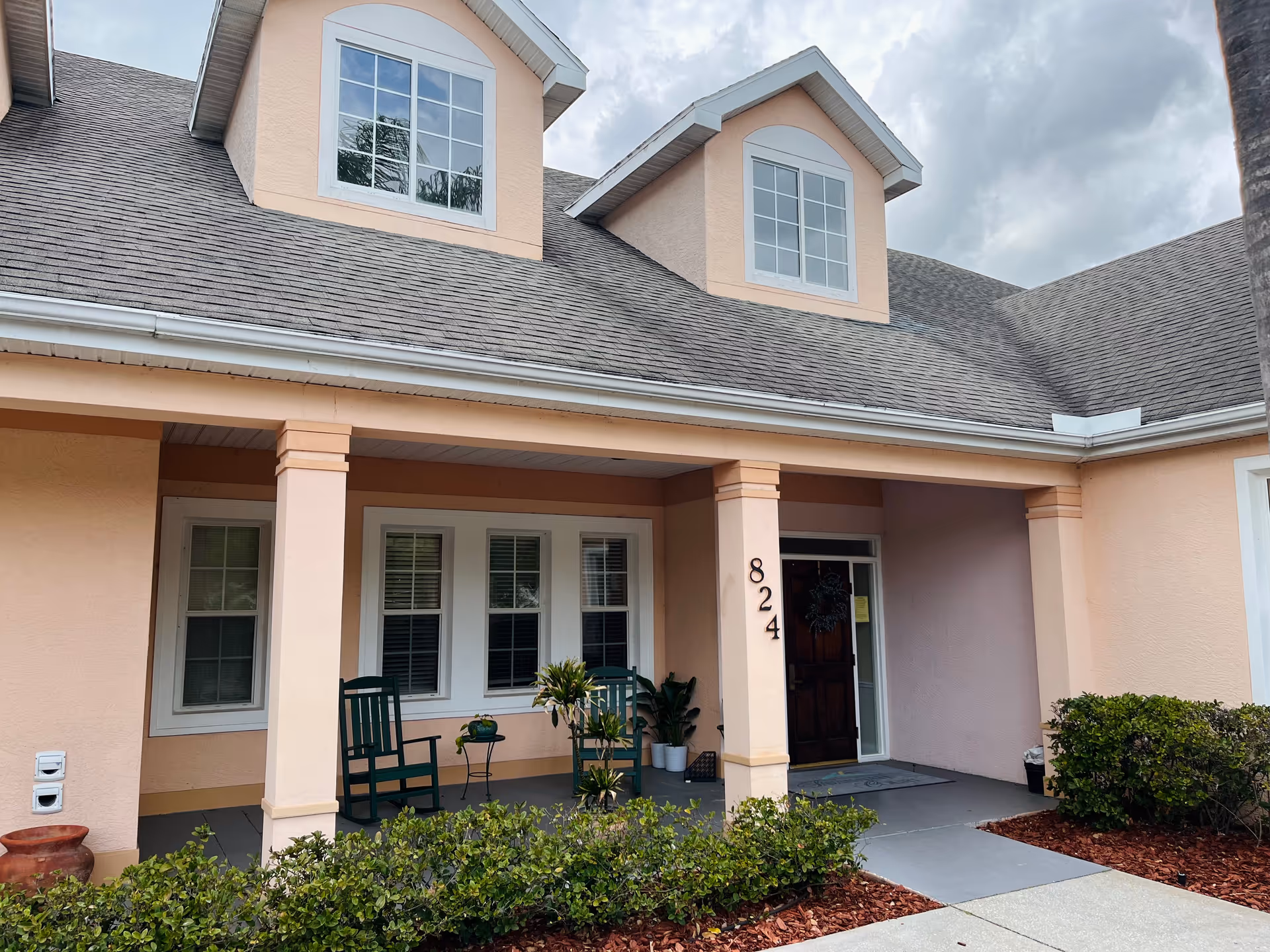 Front exterior of a peach-colored building with a covered porch, rocking chairs, potted plants, and the number 824 by the door.