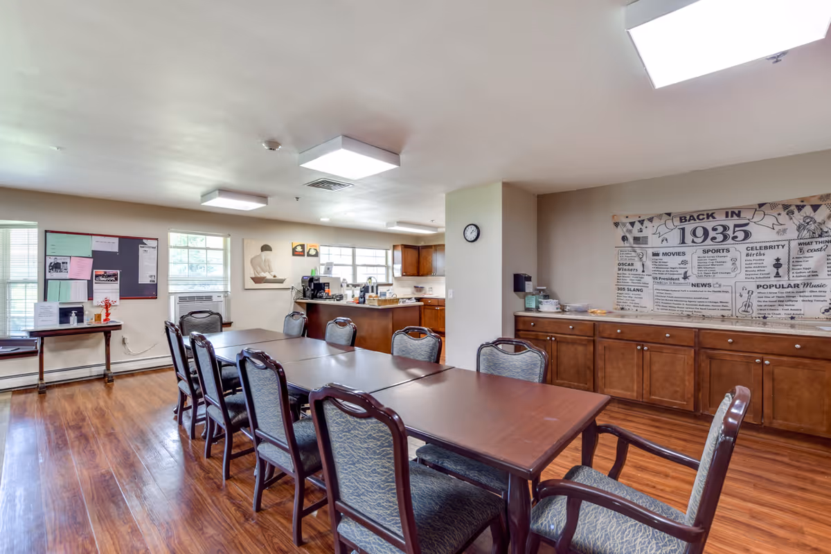 A senior living community dining room with a long wooden table surrounded by chairs. The room has wooden flooring, large windows letting in natural light, a bulletin board on the wall, and a kitchen area with cabinets and appliances in the background. There is a large decorative banner on the wall with historical information from 1935.