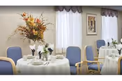 A dining room with round tables covered in white tablecloths, set with plates, glasses, and napkins. Each table has a floral centerpiece. Blue cushioned chairs with wooden armrests surround the tables. The room has large windows with sheer white curtains and valances, and a framed picture on the wall.