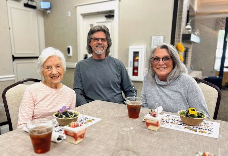 Three adults sitting at a table in a dining area, smiling at the camera. The table has cups of iced tea, small dessert cups with strawberries, and bowls with salad garnished with edible flowers. The background shows a beige wall, a door, and part of a fireplace.