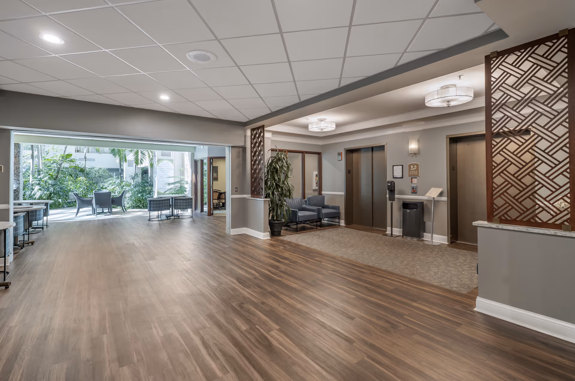 Interior view of a senior living facility hallway with wood flooring, two elevators on the right side, seating area with chairs and a potted plant, and an open area leading to a sunlit outdoor patio with chairs and greenery.