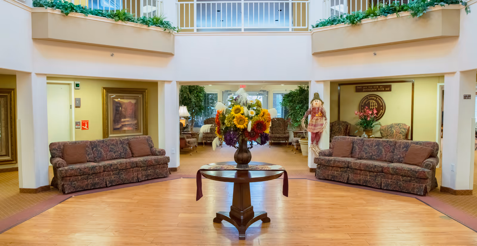 A spacious and well-lit common area in a senior living facility featuring two floral-patterned sofas facing each other with a round wooden table in the center holding a large vase of colorful flowers. The room has a high ceiling with a balcony railing above, potted plants, framed artwork on the walls, and a scarecrow decoration near one sofa. There is a sign on the wall that reads 'Don't just live with us - participate with us!'