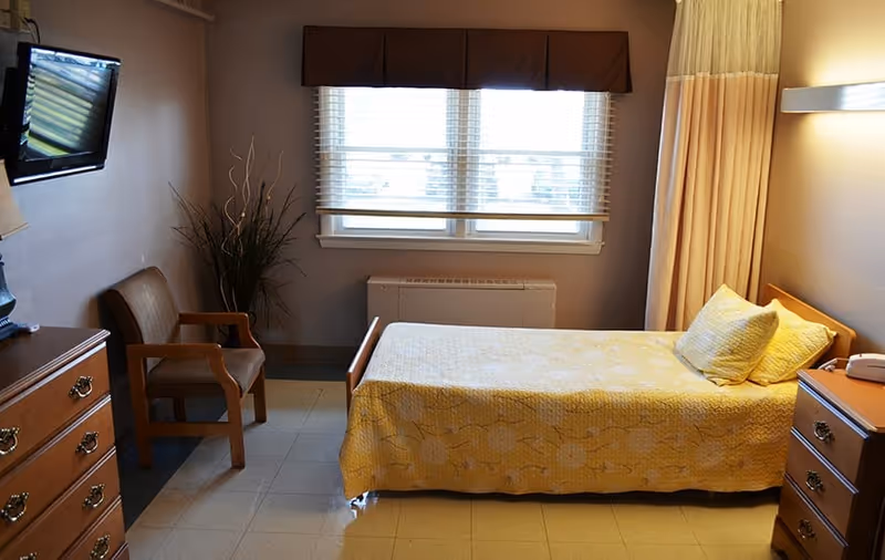 A small single-occupancy bedroom with a yellow-blanketed bed, dresser, chair, wall-mounted TV, and a window with blinds.