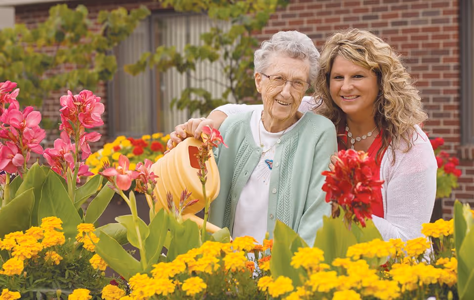 An elderly woman and a younger woman smiling together in a garden filled with colorful flowers. The elderly woman is holding a yellow watering can, and they are standing in front of a brick building with windows.