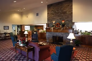 A cozy living room area in Vinecroft Retirement Community featuring a stone fireplace decorated with autumn-themed ornaments. The room has comfortable armchairs and sofas arranged around the fireplace, with side tables holding lamps and floral arrangements. The space is warmly lit with recessed ceiling lights and has a patterned carpet floor.
