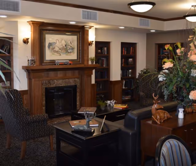 A cozy living room area with a wooden fireplace mantel adorned with a framed painting of birds. The room features built-in bookshelves filled with books, comfortable patterned armchairs, a black leather sofa, and a coffee table with decorative items. There is also a side table with a wooden deer figurine and a large floral arrangement. The lighting includes ceiling fixtures and wall sconces.