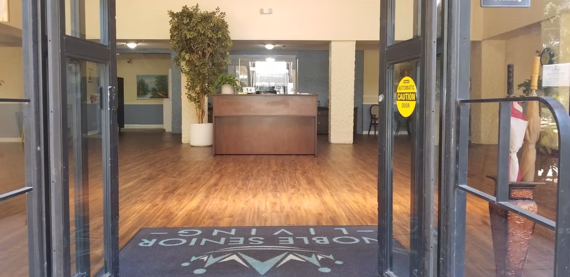 View through open glass doors into a senior living facility lobby with a wooden reception desk, a large potted plant, wood flooring, and a welcome mat on the floor. There is a yellow caution sign on the door indicating an automatic door.