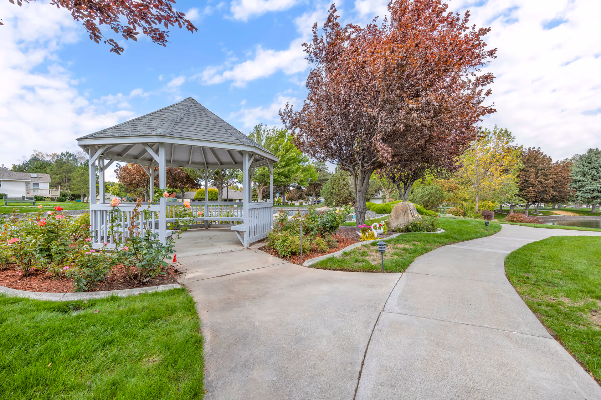 A peaceful outdoor garden area with a white gazebo surrounded by blooming rose bushes and various trees with autumn-colored leaves. A paved walkway curves around the gazebo, leading through well-maintained green lawns and landscaped flower beds under a partly cloudy sky.