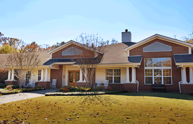 Front exterior view of a single-story brick building with white columns and a covered porch featuring rocking chairs. The building is surrounded by a well-maintained lawn with some trees and shrubs, under a clear sky.