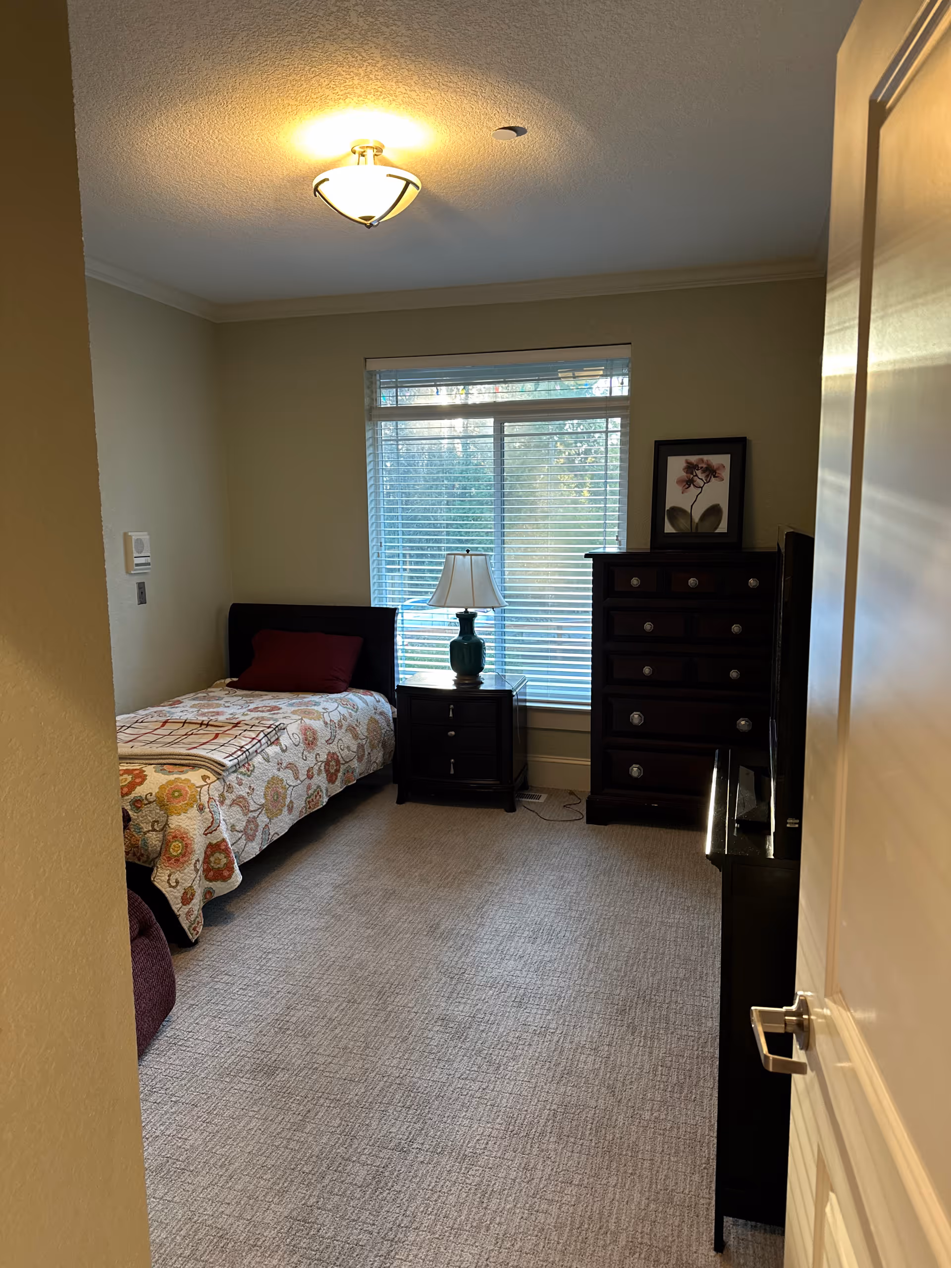 A small bedroom with a single bed covered in a floral quilt, a dark wooden nightstand with a lamp, a tall dark wooden dresser with a framed flower picture on top, and a window with blinds letting in natural light.