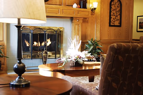 Cozy living room area with a lit fireplace surrounded by wooden paneling. A round wooden coffee table with a floral centerpiece is in front of a patterned armchair. A table lamp with a beige shade is on a side table in the foreground. Decorative wall art and plants are also visible.