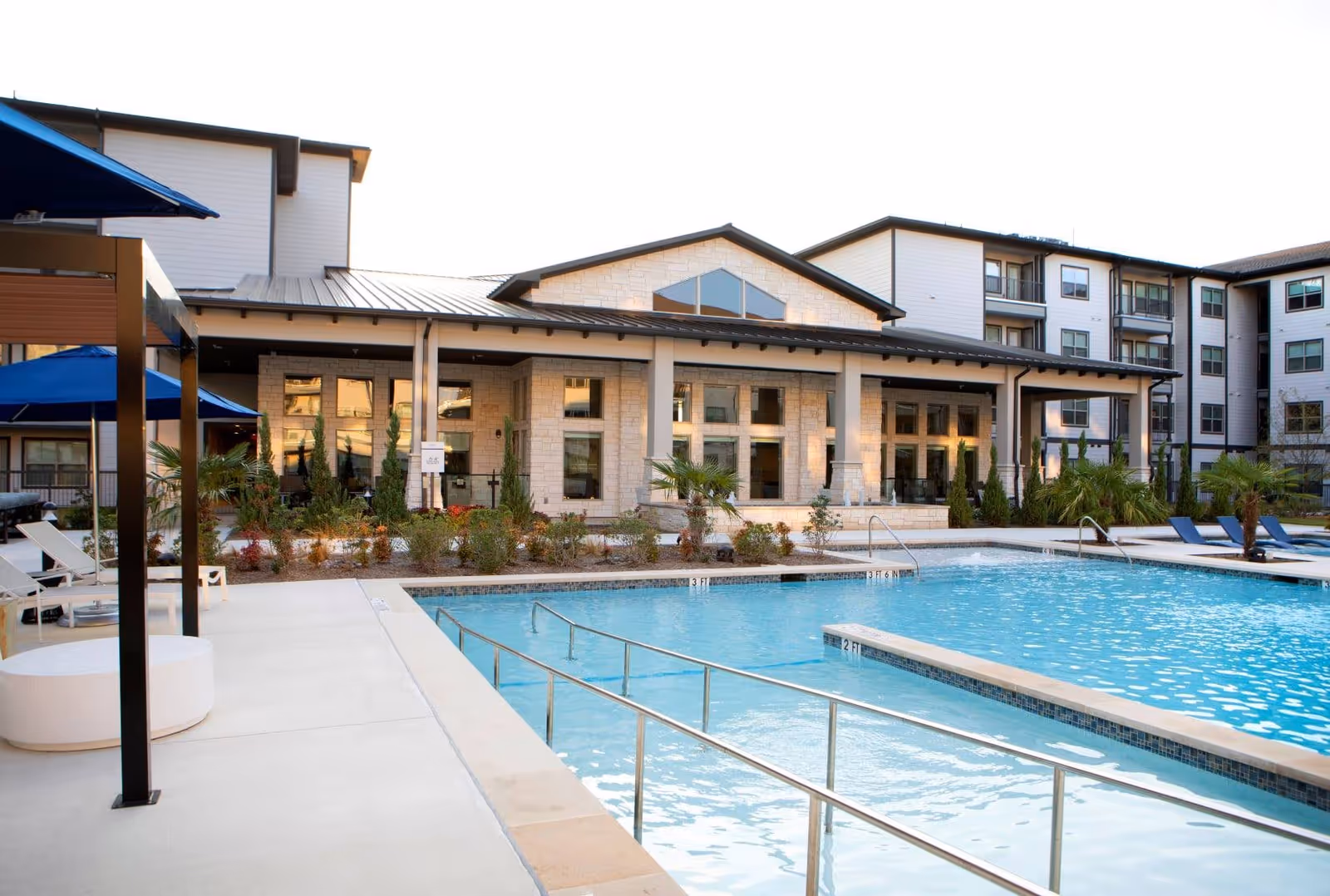 Outdoor swimming pool with handrails and lounge chairs around it, adjacent to a modern multi-story building with large windows and a covered patio area. There are plants and small trees along the edge of the building.