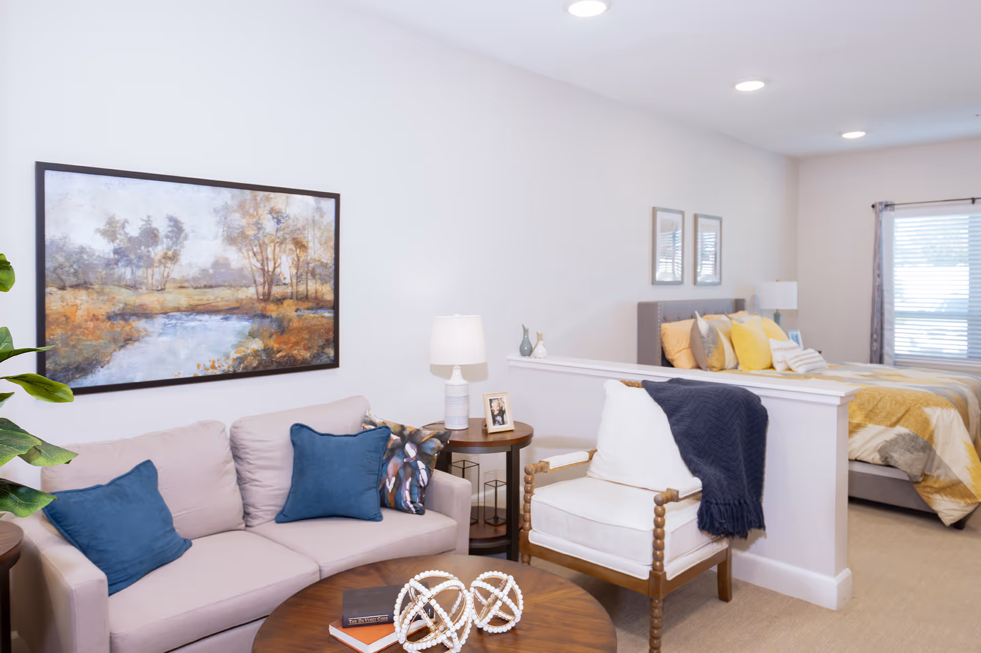 Bright open-plan living area with a sofa, armchair, coffee table and wall art, with a bed visible in the background.