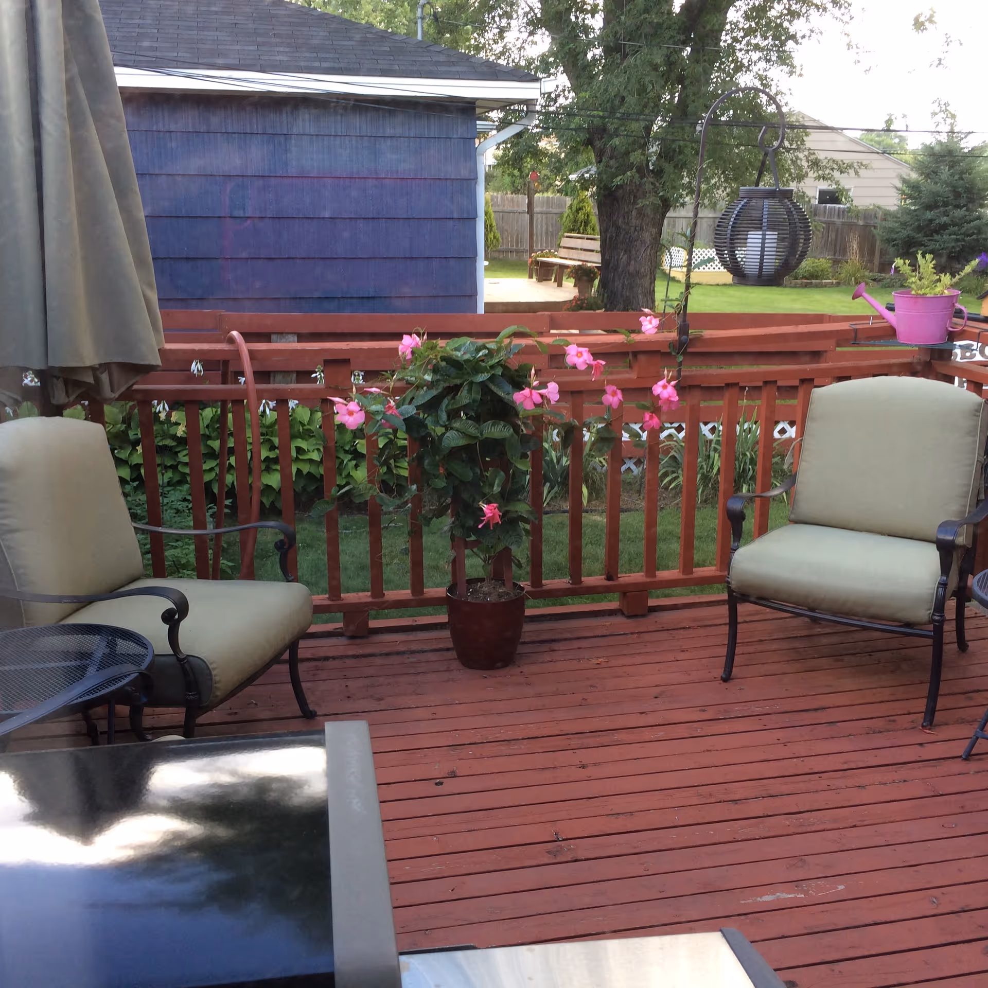 Outdoor wooden deck with two cushioned chairs, a small round metal table, a potted plant with pink flowers, a hanging lantern, and a purple watering can on the railing. In the background, there is a blue shed, a tree, a wooden bench, and a fenced yard with green grass.