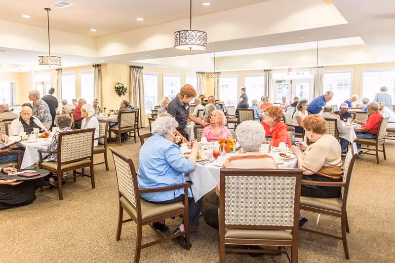A bright dining room filled with elderly residents seated at round tables covered with white tablecloths, enjoying meals and socializing. A staff member is serving or assisting one of the residents. The room has large windows with curtains, pendant lights, and a warm, welcoming atmosphere.