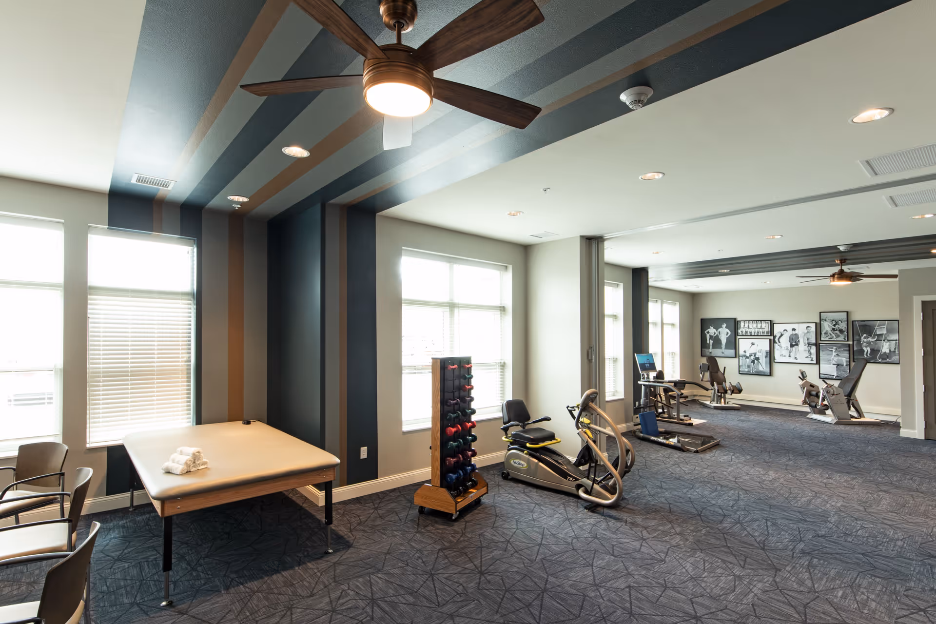 A spacious fitness room with exercise equipment including a recumbent bike, a rack of dumbbells, and other workout machines. There is a large padded table with rolled towels on it near the windows. The room has a modern design with striped ceiling and walls, ceiling fans, and framed black and white photos on the far wall.
