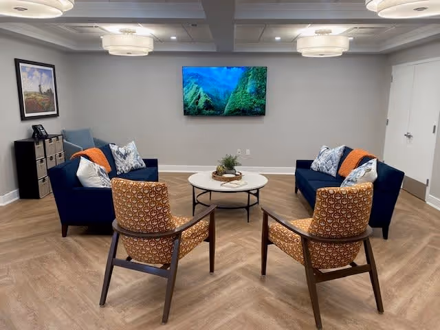A modern living room with two blue sofas adorned with patterned and solid pillows and orange throws, two patterned armchairs, a round white coffee table with decorative items, a wall-mounted TV displaying a nature scene, and ceiling lights. The room has wood-patterned flooring and light-colored walls with a framed picture and a small shelving unit in the corner.