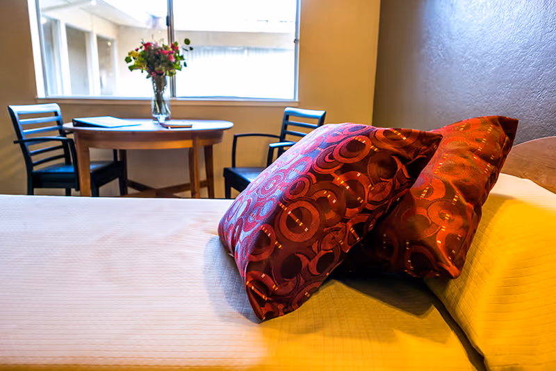 A cozy bedroom area with a bed in the foreground featuring two decorative red and orange patterned pillows. In the background, there is a round wooden table with a vase of flowers and two black chairs near a window letting in natural light.