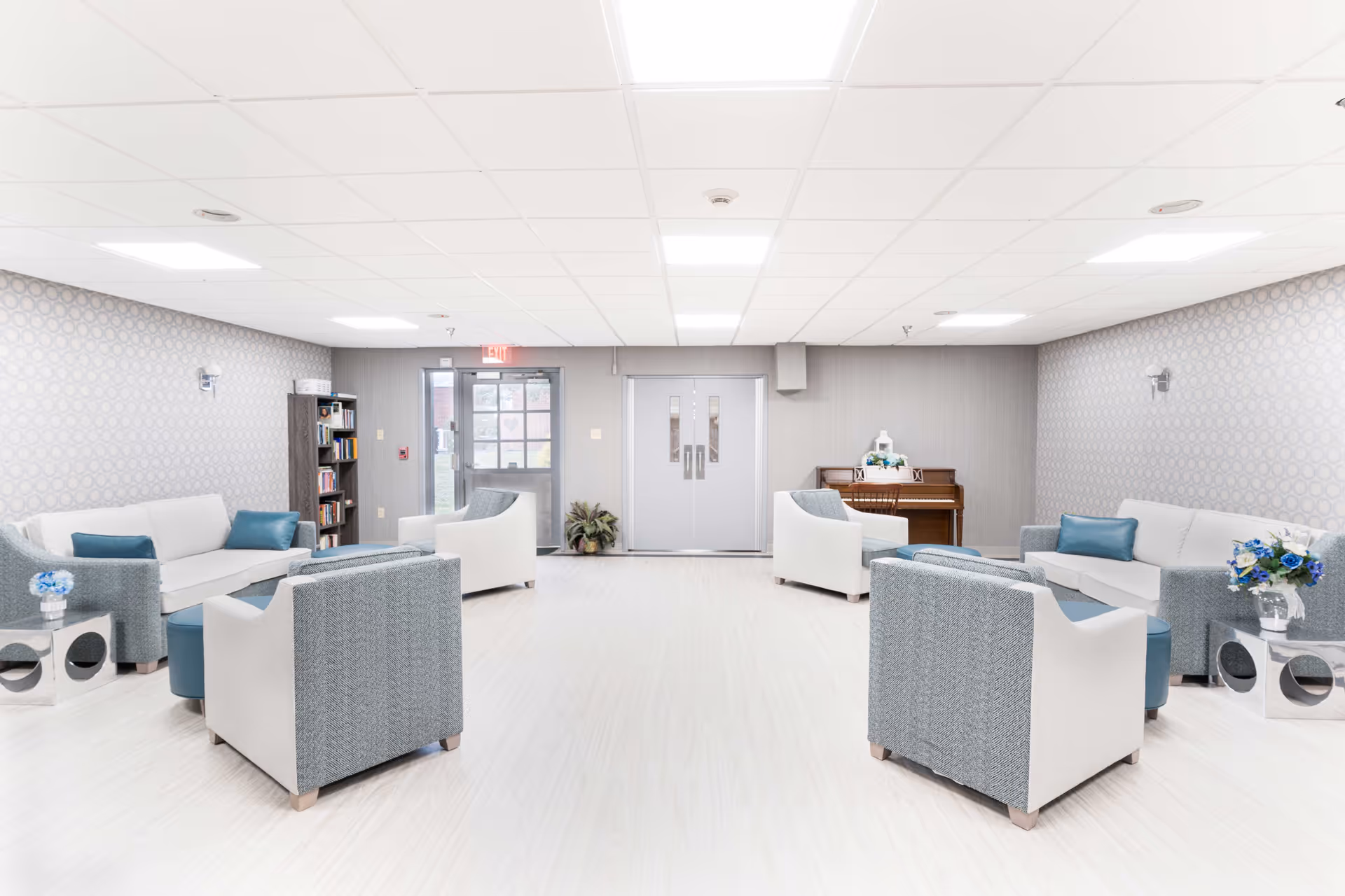 A bright and spacious common area with modern seating including sofas and armchairs arranged around small tables. The room features light-colored flooring, patterned wallpaper, and a piano against the far wall. There is a bookshelf near the exit door, and the ceiling has recessed lighting panels.