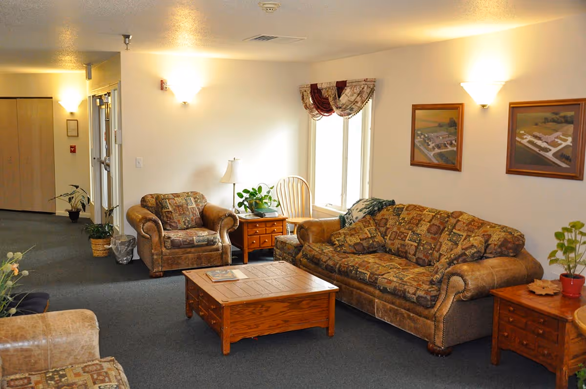 A cozy living room area with a patterned sofa and armchair, a wooden coffee table, side tables with plants and a lamp, two framed pictures on the wall, and a window with decorative curtains letting in natural light.