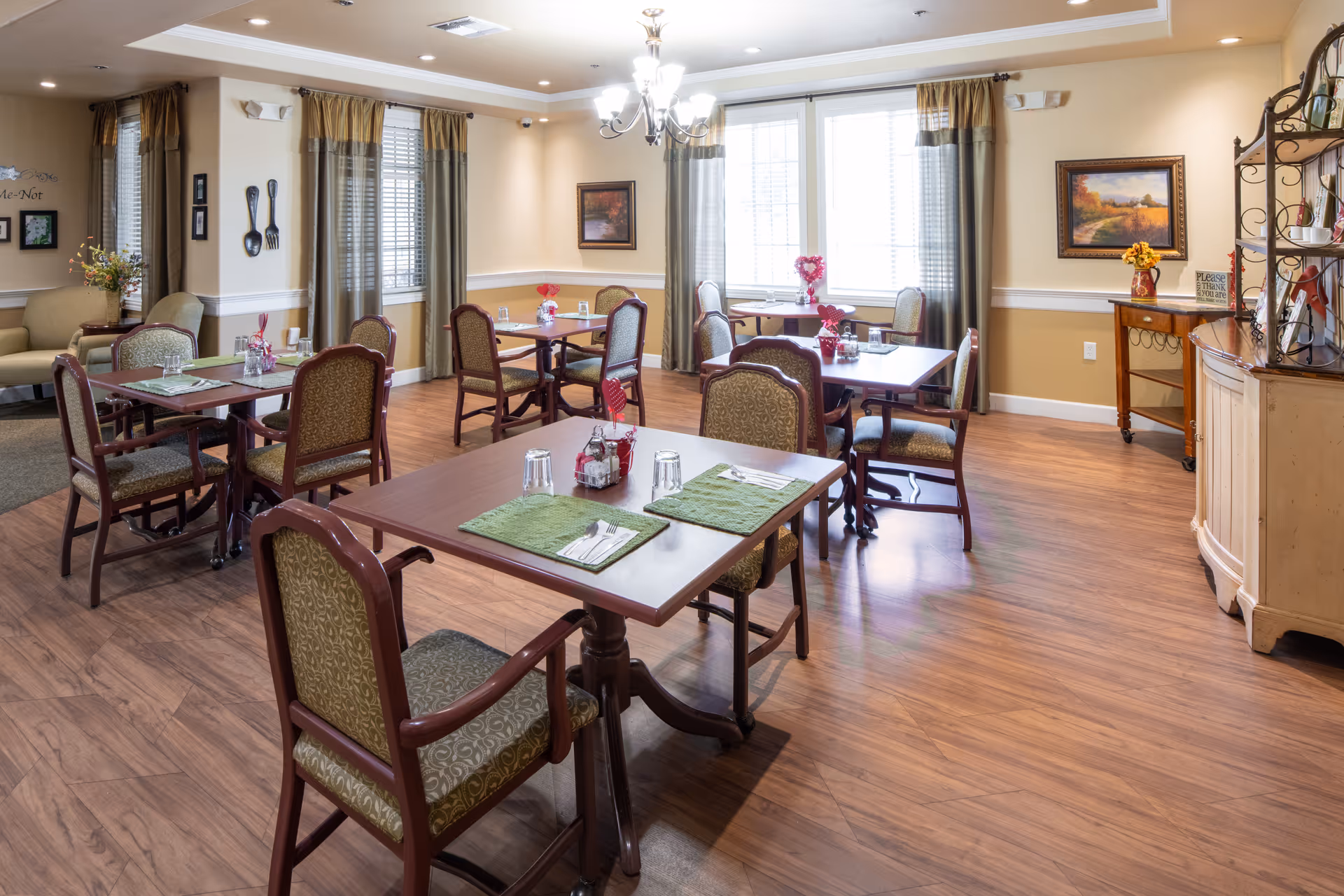 A bright and inviting dining room with several wooden tables and cushioned chairs arranged neatly. Each table is set with green placemats, glasses, and silverware. The room features wood flooring, large windows with curtains allowing natural light, and warm beige walls adorned with framed artwork and decorative items. A chandelier hangs from the ceiling, and there is a sideboard with flowers and decor on the right side.