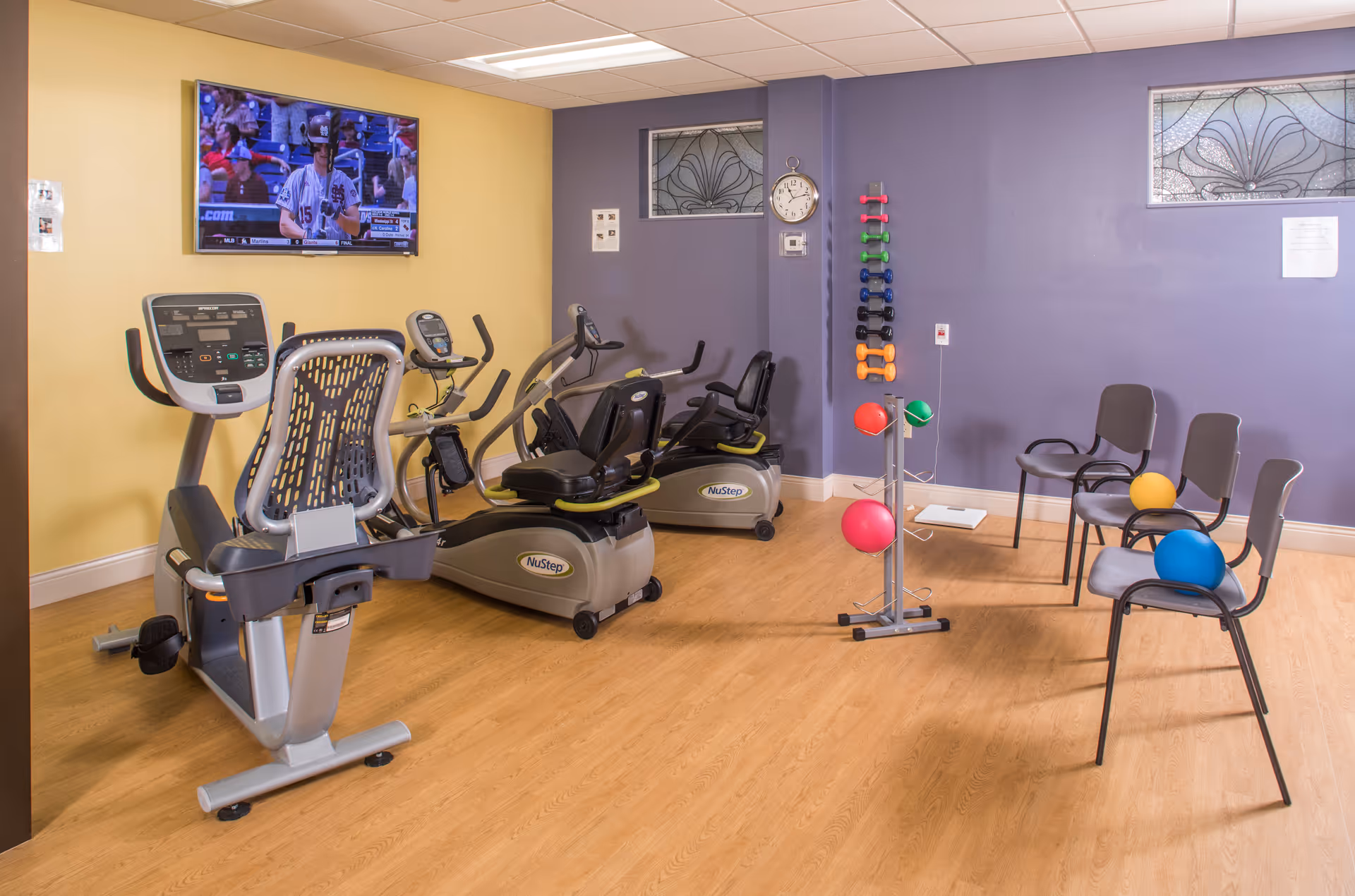 A small exercise room with two NuStep recumbent exercise bikes, a rack of colorful dumbbells, three chairs each holding a different colored exercise ball, a wall clock, and a wall-mounted TV showing a baseball game. The walls are painted yellow and purple with decorative windows near the ceiling.