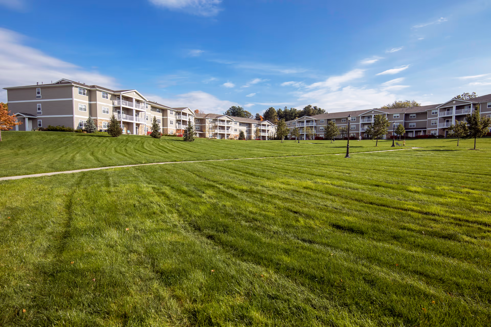 Wide view of a senior living facility with multiple three-story buildings surrounding a large, well-maintained grassy lawn with a paved walking path and several small trees under a partly cloudy blue sky.