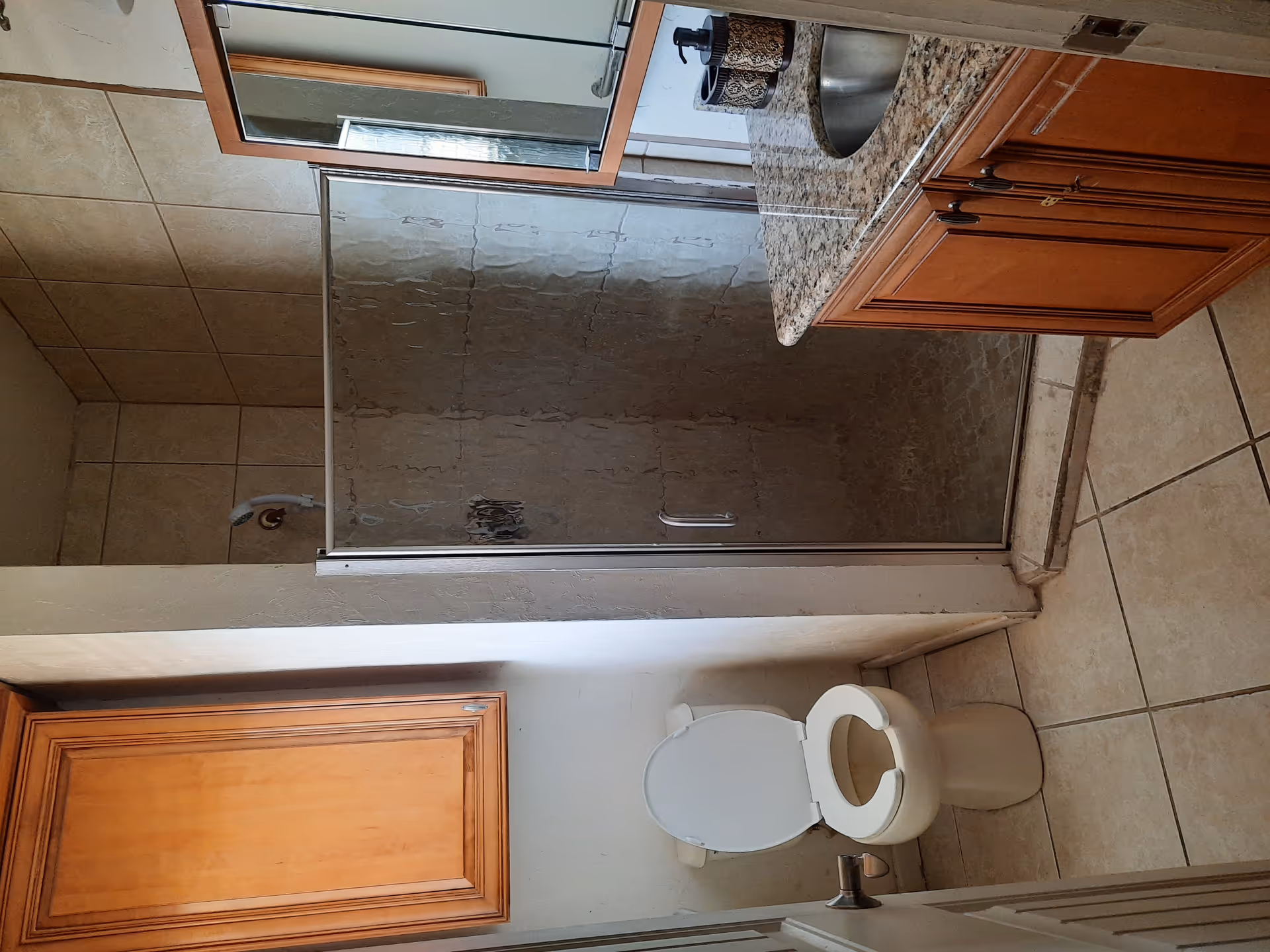 A small bathroom with beige tiled floor and walls. There is a white toilet with the seat lid open on the left side, a wooden cabinet above it, a glass-enclosed shower with a silver frame in the center, and a wooden vanity with a granite countertop and a round sink on the right. A mirror with a wooden frame is mounted above the sink.