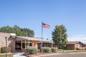 Single-story brick building with large windows and an American flag on a flagpole in front, surrounded by trees and landscaping under a clear blue sky.