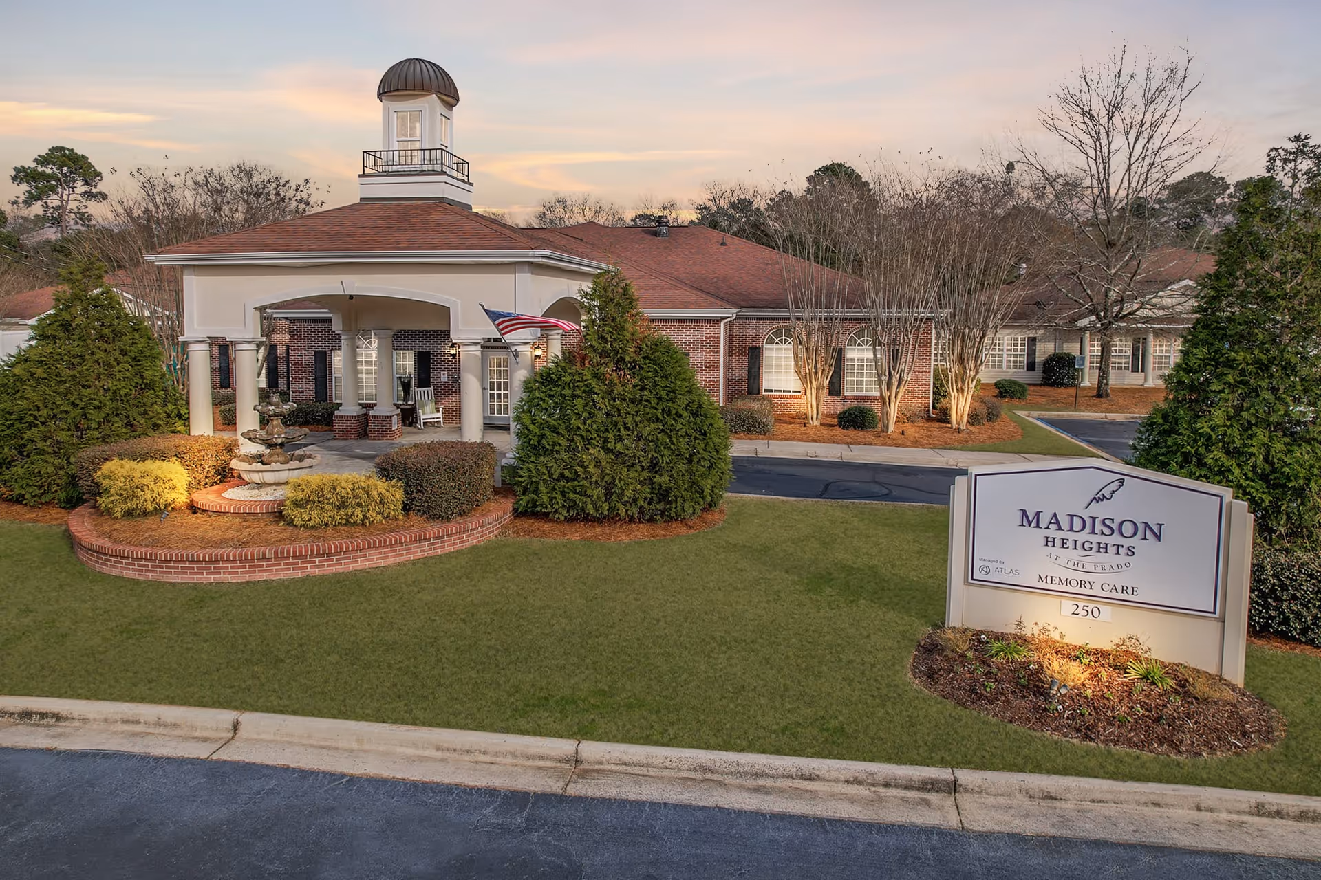 Exterior view of a senior living facility named Madison Heights at The Prado Memory Care. The building has a brick facade with white columns supporting a covered entrance topped by a small cupola. There is a landscaped area with bushes and a fountain in front, and a sign displaying the facility name and address number 250. The sky is partly cloudy with a warm light suggesting early evening.