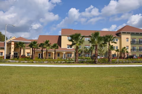Exterior view of a senior living facility building with a beige and green facade, red roofs, and several palm trees in front. The sky is partly cloudy and there is a well-maintained lawn in the foreground.