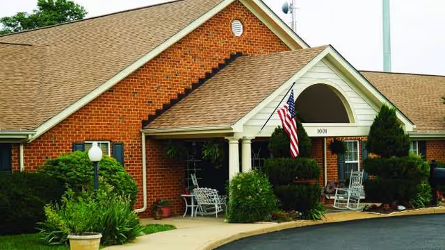 Brick senior living facility front with arched entrance, flag, and front porch seating.