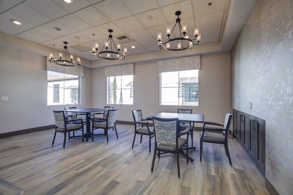 A bright dining room with three square tables, each surrounded by four chairs with patterned upholstery. The room has large windows with patterned valances, wood flooring, and three modern chandeliers hanging from a white ceiling. There is a textured accent wall with built-in dark wood cabinets.