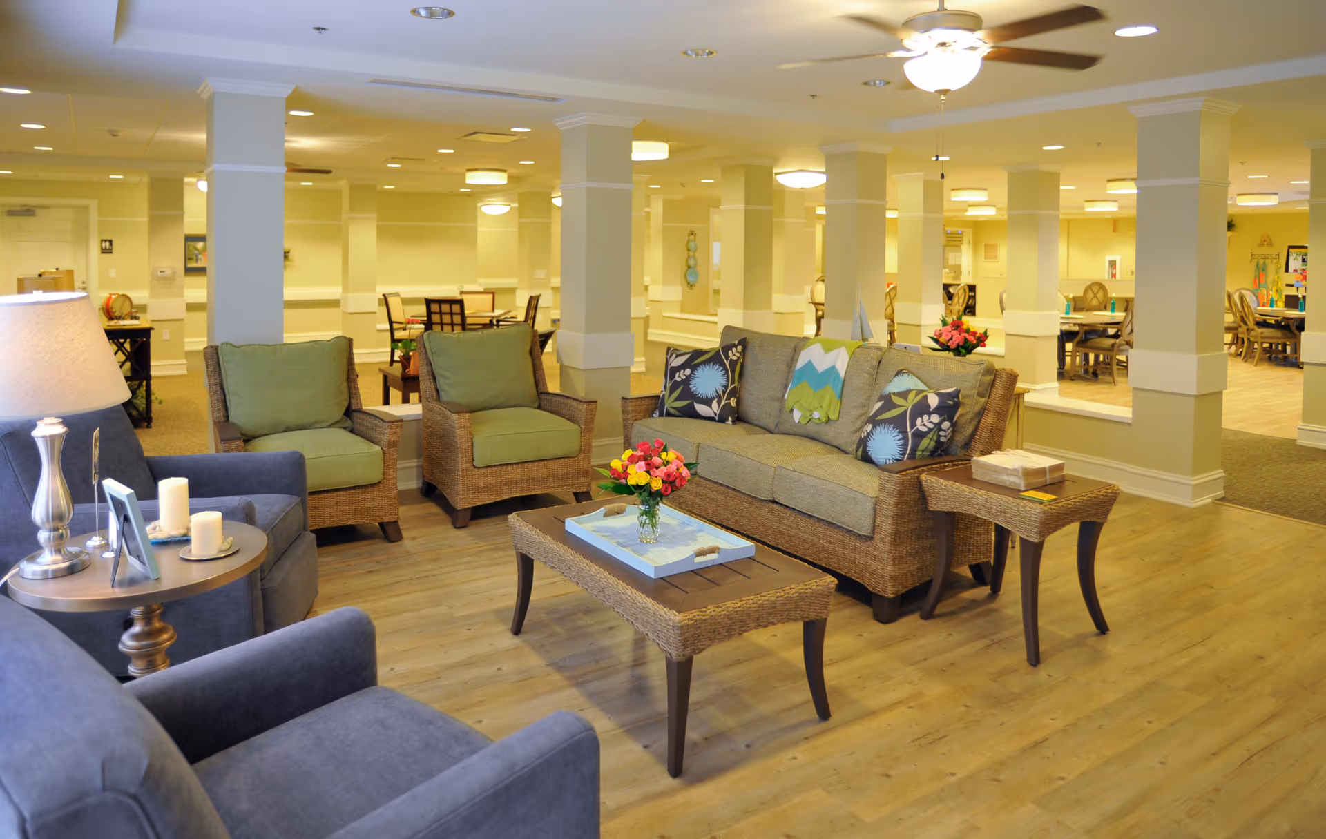 A bright and spacious senior living common area with comfortable seating including a sofa with decorative pillows, two armchairs, and two single chairs. The room features wooden flooring, several columns, and multiple tables with flowers and candles. In the background, there are more tables and chairs arranged for dining or activities under warm ceiling lights.