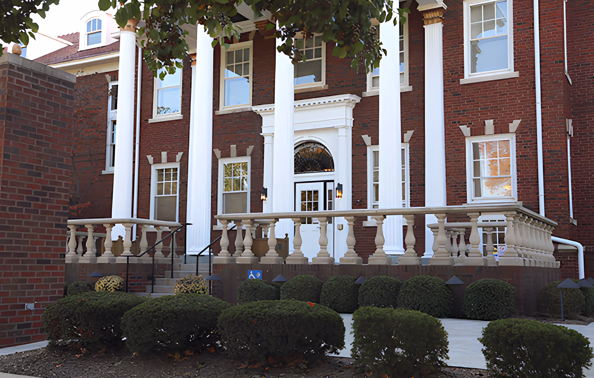 Front exterior view of a red brick building with large white columns and a white door entrance. There are multiple windows, trimmed bushes, and a small ramp with handrails leading to the entrance. The building has a classic architectural style with a porch and balustrade.