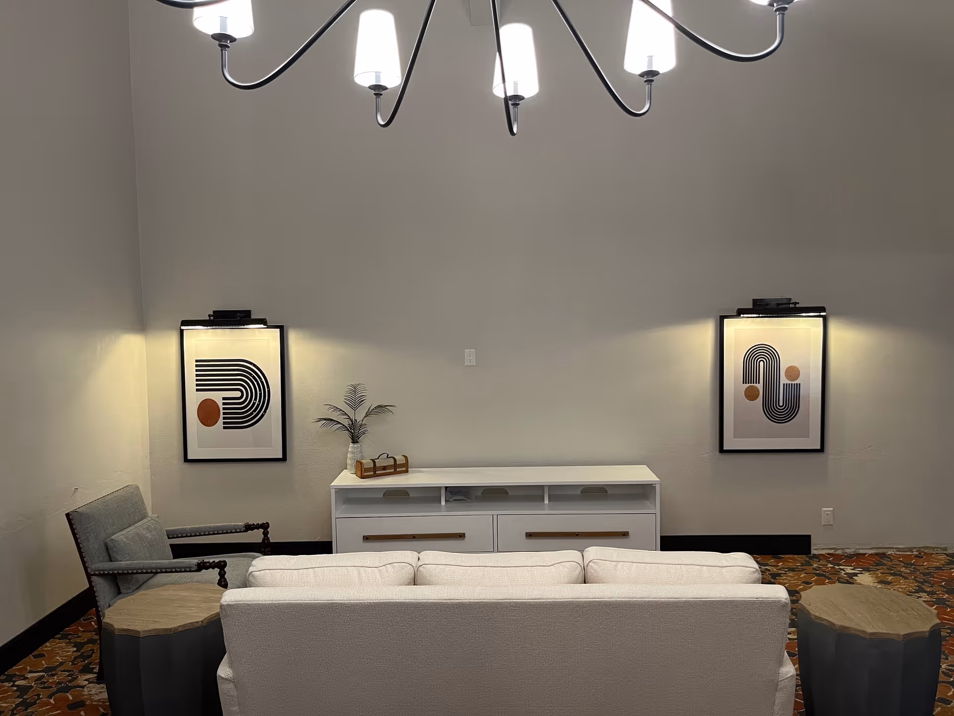 A modern sitting area with a white sofa facing a console table, flanked by an armchair, two framed abstract prints, and an overhead chandelier.
