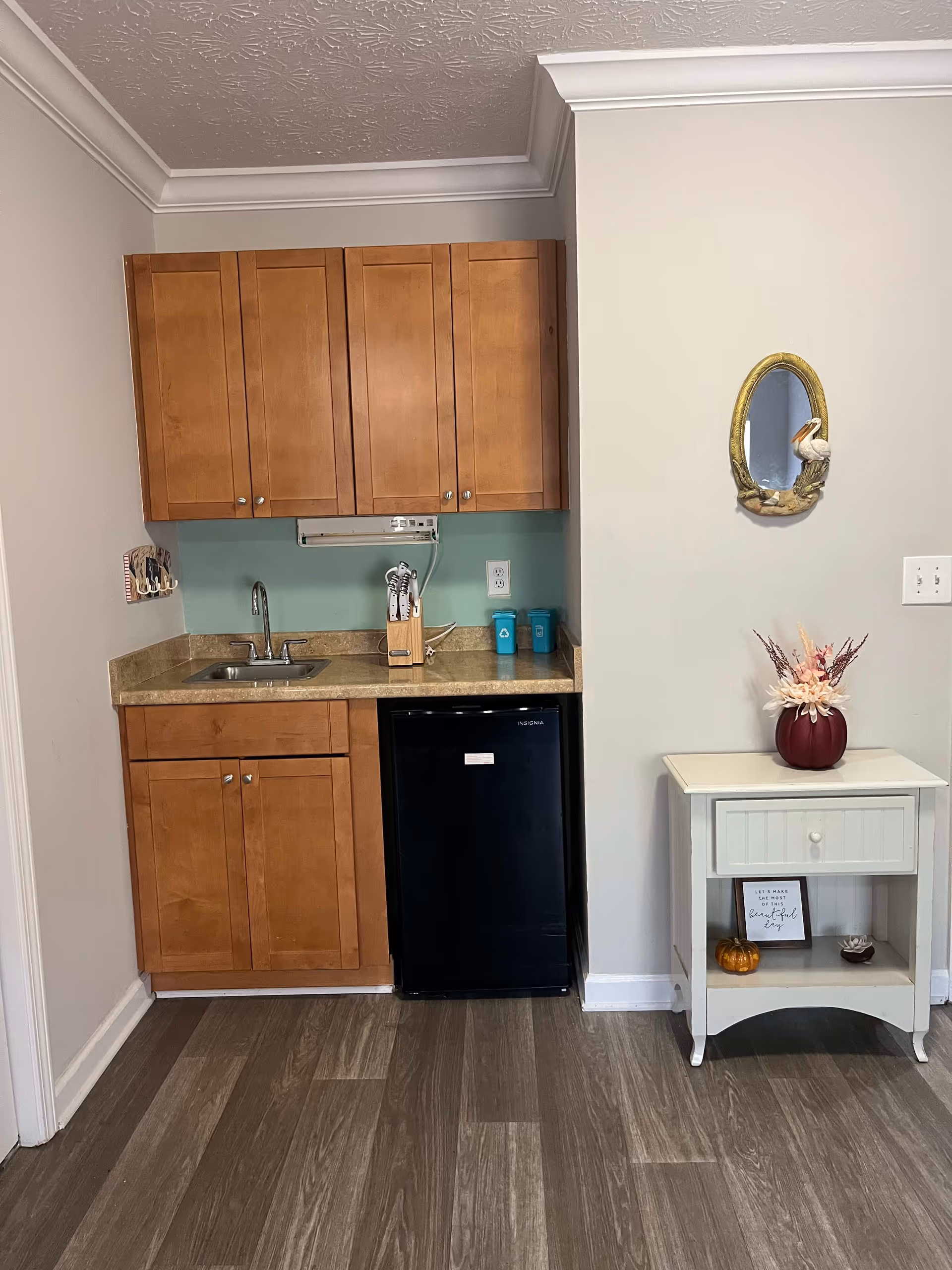 Small kitchenette area with wooden cabinets, a countertop with a sink, a knife block, and a small black refrigerator. To the right, there is a white side table with a drawer and an open shelf, decorated with a small pumpkin, a framed quote, and a vase with flowers. Above the table, an oval mirror with a pelican decoration is mounted on the wall. The floor has wood-patterned flooring and the walls are painted light gray.
