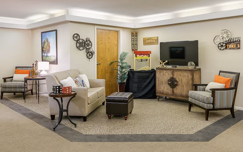 A cozy living room area with a beige loveseat, two striped armchairs with orange and white pillows, a small round side table with decorative dice, a wooden cabinet with a flat-screen TV on top, a popcorn machine, and wall decorations including film reels and theater signs.