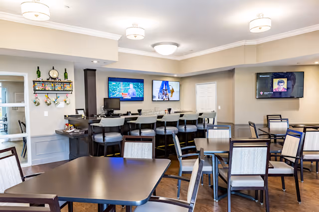 A modern dining area in a senior living facility featuring multiple tables and chairs arranged neatly. There is a bar counter with several high chairs, and three flat-screen TVs mounted on the walls. The room is well-lit with ceiling lights and has a neutral color scheme.