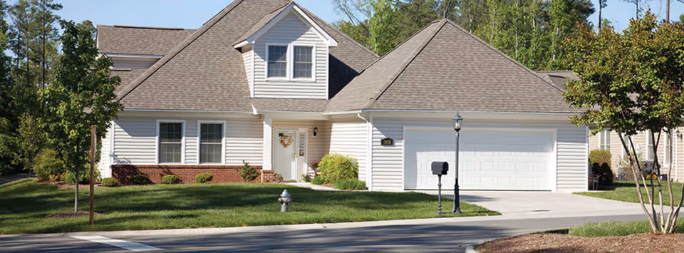 A single-story house with white siding and a gray shingled roof, featuring a front door with a decorative wreath, a two-car garage, a mailbox, a street lamp, and a well-maintained lawn with small trees and shrubs.